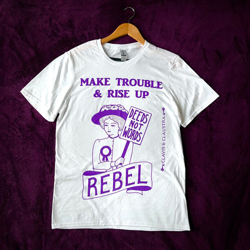 Photo of a white t-shirt with an illustration and text, all in purple. The image is of a suffragette wearing a hat with flowers, a rosette, and holding a sign that says “Deeds Not Words”. Beneath her is a banner that says “Rebel”. Above is the caption “Make Trouble & Rise Up”.