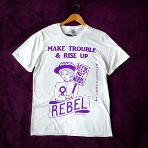 Photo of a white t-shirt with an illustration and text, all in purple. The image is of a suffragette wearing a hat with flowers, a rosette, and holding a sign that says “Deeds Not Words”. Beneath her is a banner that says “Rebel”. Above is the caption “Make Trouble & Rise Up”.