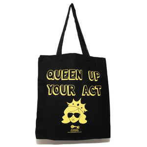 Photo of a black tote bag. The bag says “Queen Up Your Act” in yellow outline letters and features the image of an unsmiling woman with long hair wearing sunglasses and a crown. The handles are up in the air.