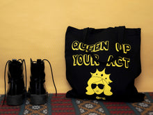 Load image into Gallery viewer, Photo of a black tote bag resting against the wall on a carpet. The bag says “Queen Up Your Act” in yellow outline letters and features the image of an unsmiling woman with long hair wearing sunglasses and a crown. Next to the bag stands a pair of tall black boots.