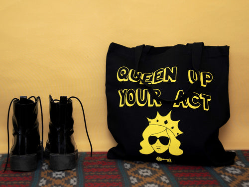 Photo of a black tote bag resting against the wall on a carpet. The bag says “Queen Up Your Act” in yellow outline letters and features the image of an unsmiling woman with long hair wearing sunglasses and a crown. Next to the bag stands a pair of tall black boots.
