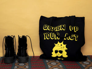 Photo of a black tote bag resting against the wall on a carpet. The bag says “Queen Up Your Act” in yellow outline letters and features the image of an unsmiling woman with long hair wearing sunglasses and a crown. Next to the bag stands a pair of tall black boots.
