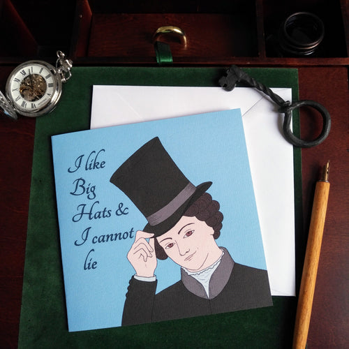 Photo of a blue greeting card with an illustration of Anne Lister, a white woman wearing a black jacket and top hat which she tips to the viewer with a smile. The caption reads “I like Big Hats and I cannot lie.” The card is on a brown desk and white envelope, surrounded by an iron key, a silver pocket watch, and a yellow pencil.