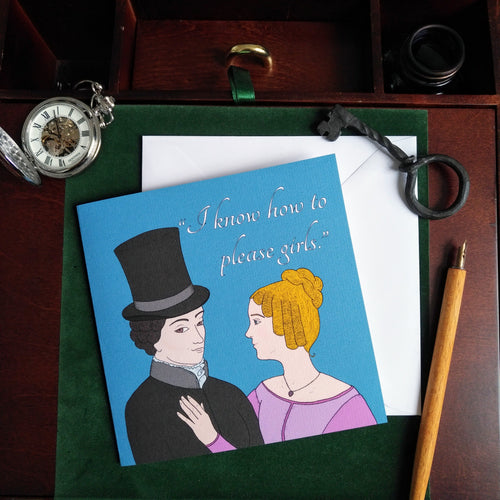 Photo of a blue greeting card with an illustration of Anne Lister, a white woman wearing a top hat and black jacket and looking at the viewer with a smile, alongside Ann Walker, a white woman with blonde hair and a pink dress, who is resting her hand on Anne Lister’s chest and looking to her. The caption reads “I know how to please girls.” The card is on a brown desk and white envelope, surrounded by an iron key, a silver pocket watch, and a yellow pencil.
