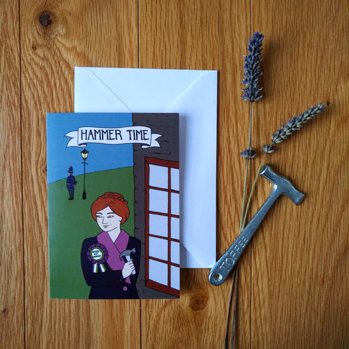 Photo of a greeting card depicting a white female suffragette with brown hair and a “Votes for Women” rosette holding a hammer next to a window while she looks at an unaware policeman and smirks. The caption reads “Hammer Time.” Next to the card is a white envelope, replica toffee hammer, and a sprig of lavender.