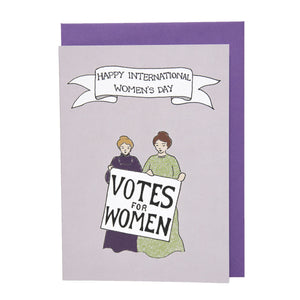 Photo of a greetings card depicting two suffragettes standing together and holding a sign that reads “Votes For Women”. Above is a banner reading "Happy International Women’s Day". Also pictured is the card’s purple envelope.
