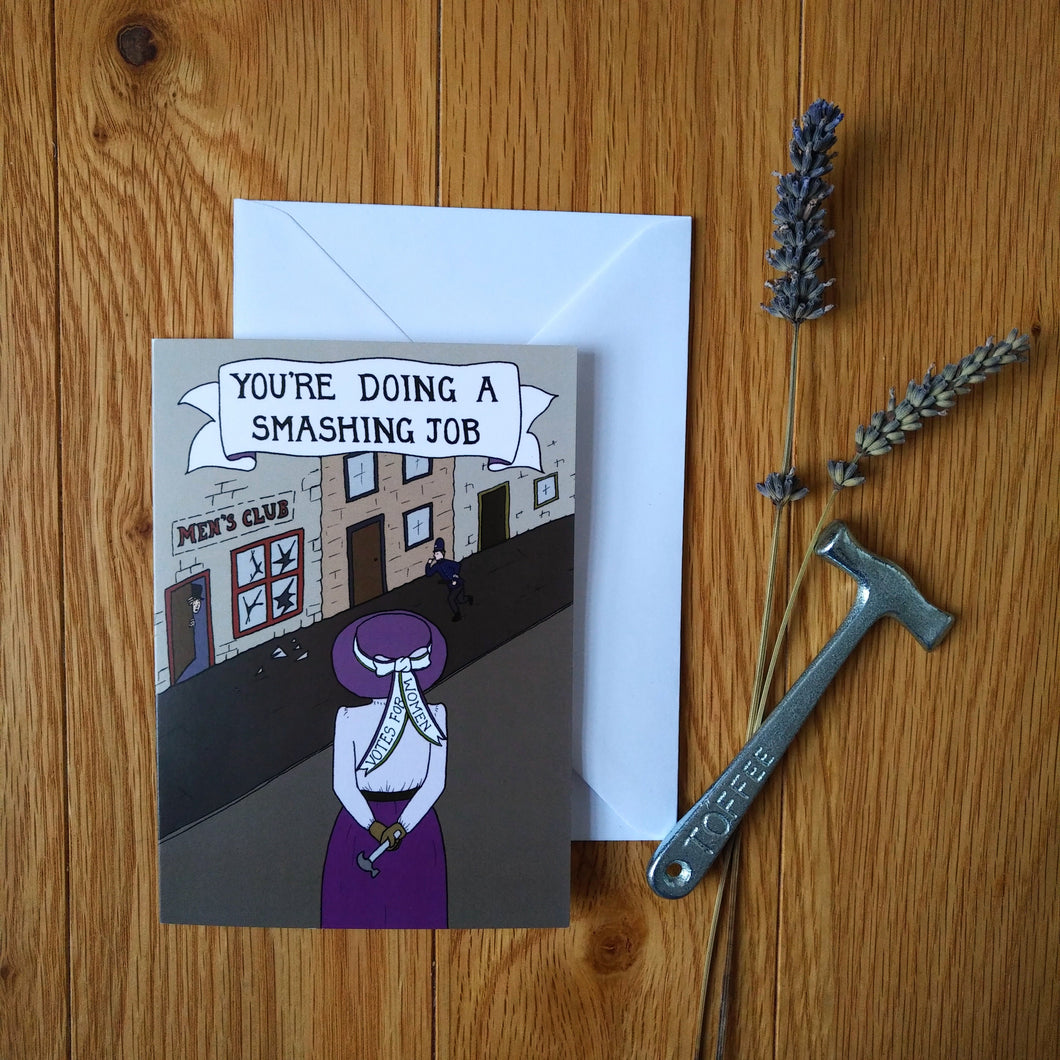 Photo of the card which shows the back of a suffragette in a purple hat, shirt and skirt as she holds a toffee hammer behind her. She surveys a Men’s Club where the windows have all been broken. A man meekly peeks from out the cracked door. The banner caption reads “You’re Doing A Smashing Job”. Next to the card is a white envelope, some lavender, and a toffee hammer.