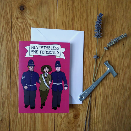 Photo of the dark pink greeting card. The artwork shows a suffragette in a “Votes For Women” sash being led away in handcuffs by two policemen in uniform while she shouts. The caption reads “Nevertheless She Persisted” on a banner. Next to the card is some lavender and a toffee hammer.