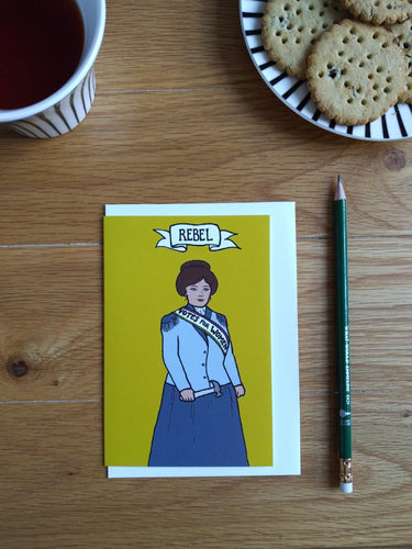 Photo of a mustard-coloured card laid on a white envelope on a table. The card shows a suffragette in a blue blazer and darker blue skit. She wears a sash that says “Votes For Women” and holds a toffee hammer in her hand. The banner at the top reads “Rebel”. Next to the card is a pencil, a mug of black tea, and a plate with biscuits on.