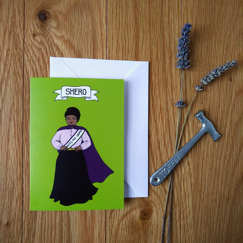 Photo of a green card with a suffragette with hands on her hips wearing a purple cloak and a sash that says “Votes For Women”. The card is next to a sprig of lavender and a toffee hammer.