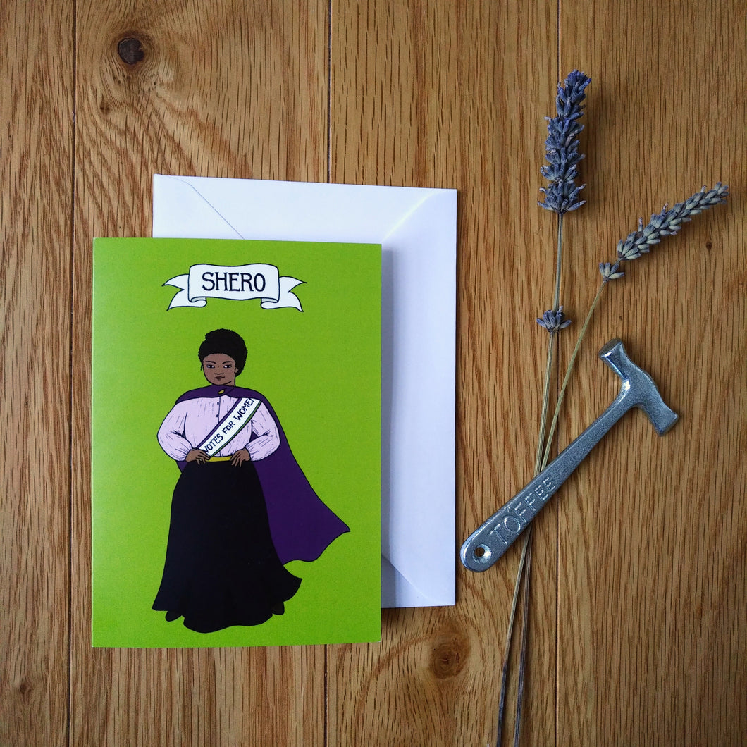 Photo of a green card with a suffragette with hands on her hips wearing a purple cloak and a sash that says “Votes For Women”. The card is next to a sprig of lavender and a toffee hammer.