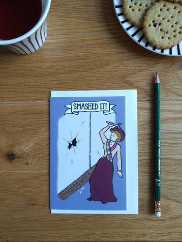 Photo of a purple greetings card. The image shows a suffragette wearing a “Votes For Women” sash and red hat wielding a toffee hammer. She stands next to a large window with a giant break in the middle and cracks emerging from the centre. Broken glass is on the floor. The banner caption reads “Smashed It!”. Next to the card is a green pencil, a mug of black tea, and a plate with biscuits on.