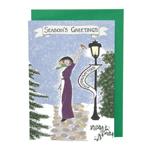 Photo of a card showing Emily Davison in a purple and white dress with a black hood. She stands on a path surrounded by snow and an old-fashioned lamppost. She has a raised arm, with a robin at rest on her fingers. From the lamppost is a banner that says “#VOTINGMATTERS”. The main caption says “Season’s Greetings” on a banner. The card is on top of a green envelope.