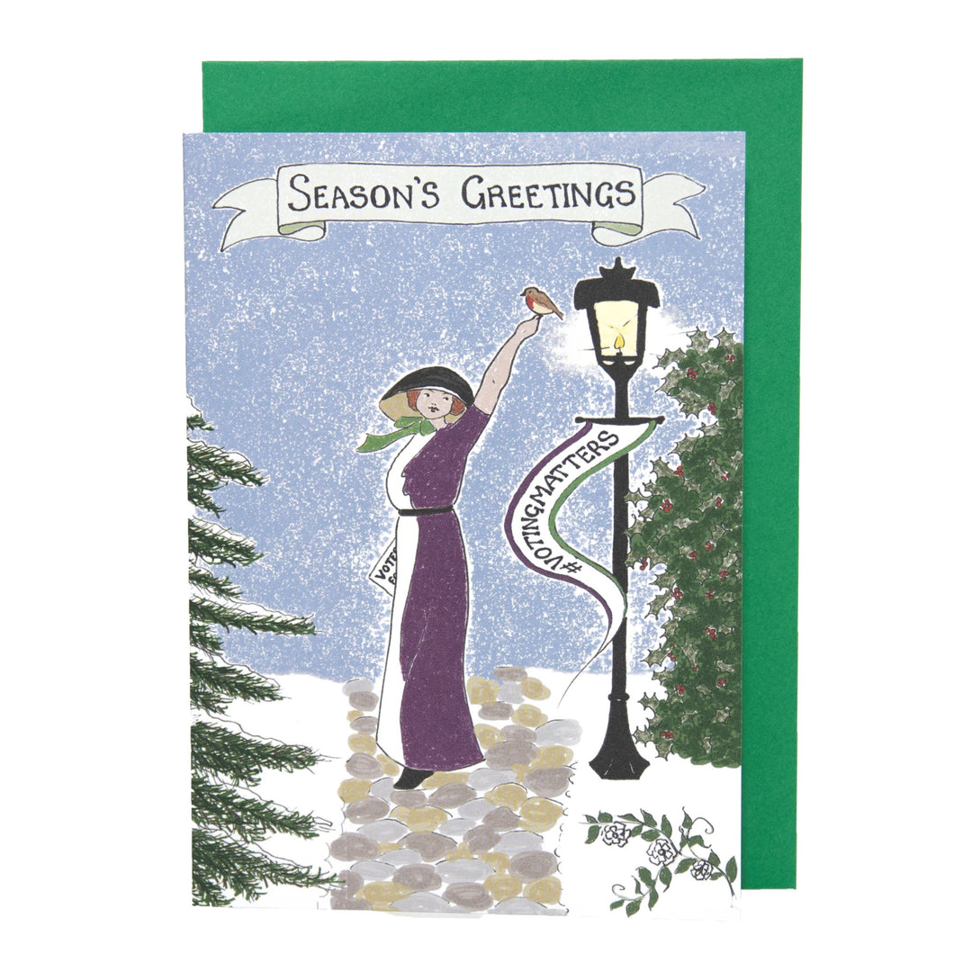 Photo of a card showing Emily Davison in a purple and white dress with a black hood. She stands on a path surrounded by snow and an old-fashioned lamppost. She has a raised arm, with a robin at rest on her fingers. From the lamppost is a banner that says “#VOTINGMATTERS”. The main caption says “Season’s Greetings” on a banner. The card is on top of a green envelope.