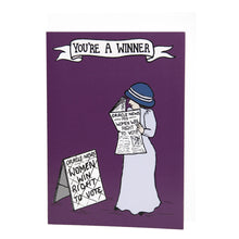 Load image into Gallery viewer, Photo of the card, mostly purple, showing a suffragette in a long dress and hat as she smiles and reads a newspaper called “Oracle News” with the front-page text “-1918- Women Win Right To Vote”. She stands next to a news board that says the same. The banner caption says “You’re A Winner!”.