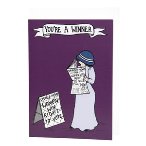 Photo of the card, mostly purple, showing a suffragette in a long dress and hat as she smiles and reads a newspaper called “Oracle News” with the front-page text “-1918- Women Win Right To Vote”. She stands next to a news board that says the same. The banner caption says “You’re A Winner!”.