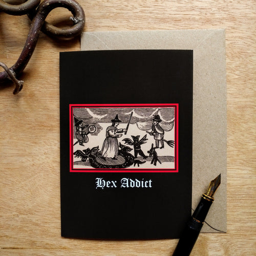 Photo of a black coloured card featuring a mediaeval woodcut artwork depicting a witch in a circle holding a wand, surrounded by a rabbit walking on its hind legs and a small demon. Two witches ride broomsticks in the sky. Beneath it the caption reads 'Hex Addict'. Pictured with a brown envelope and a black fountain pen and a curved piece of wood.
