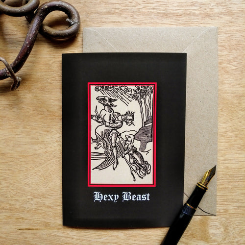 Photo of a card featuring a black-and-white mediaeval woodcut artwork on a black background, depicting three creatures with human bodies but the heads of a donkey, bird, and a dog all clinging to a flying broomstick as winds blow in the background against the trees. Beneath it the caption reads 'Hexy Beast' in a white Gothic typeface. Next to the card is a brown envelope, black fountain pen, and curved piece of wood.
