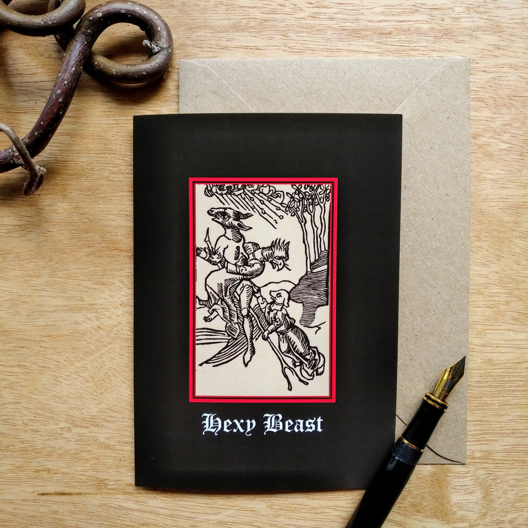 Photo of a card featuring a black-and-white mediaeval woodcut artwork on a black background, depicting three creatures with human bodies but the heads of a donkey, bird, and a dog all clinging to a flying broomstick as winds blow in the background against the trees. Beneath it the caption reads 'Hexy Beast' in a white Gothic typeface. Next to the card is a brown envelope, black fountain pen, and curved piece of wood.
