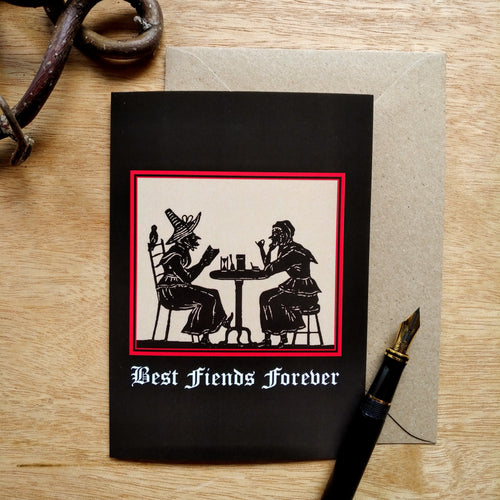 Photo of a greetings card featuring an early modern woodcut image of two witch-like women socializing over a small table. Underneath is the caption 'Best Fiends Forever' and the woodcut is surrounded by a scarlet border and the background is black. Along with the brown envelope, also pictured are a black fountain pen and a gnarled branch.