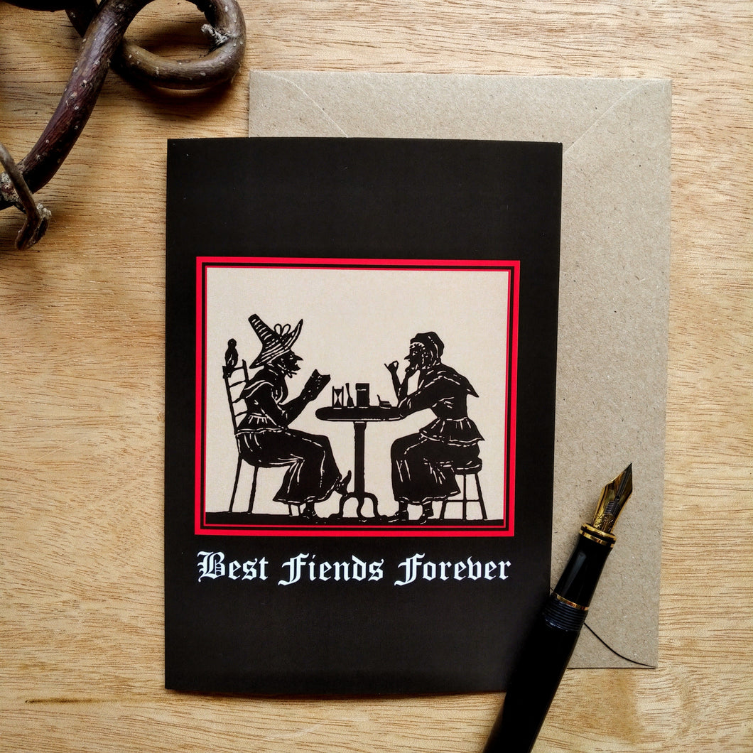 Photo of a greetings card featuring an early modern woodcut image of two witch-like women socializing over a small table. Underneath is the caption 'Best Fiends Forever' and the woodcut is surrounded by a scarlet border and the background is black. Along with the brown envelope, also pictured are a black fountain pen and a gnarled branch.