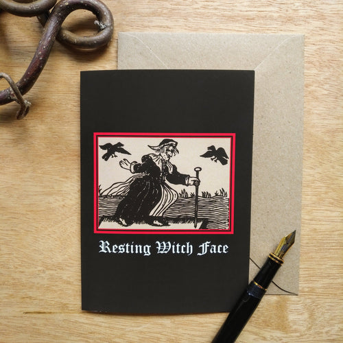Photo of a black card with a black-and-white mediaeval woodcut artwork on a black background and framed in red, depicting a stereotypical crone with warts and a large nose in a black dress, holding a cane and riding a raft on water. Two black birds hover in the sky. The caption reads 'Resting Witch Face' in a white Gothic typeface. Next to the card is a brown envelope, black fountain pen, and a twisted piece of wood.