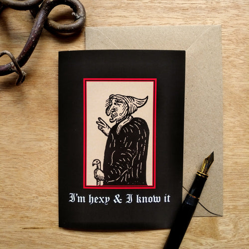 Photo of a card featuring a black-and-white mediaeval woodcut artwork on a black background, depicting a stereotypical crone with a large nose, curved chin, and a warts. She has a pointed hat and cane and holds up two fingers like scissors. Beneath it the caption reads 'I’m hexy & I know it' in a white Gothic typeface. Next to the card is a brown envelope, black fountain pen, and curved piece of wood.