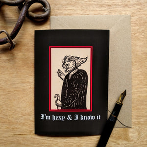 Photo of a card featuring a black-and-white mediaeval woodcut artwork on a black background, depicting a stereotypical crone with a large nose, curved chin, and a warts. She has a pointed hat and cane and holds up two fingers like scissors. Beneath it the caption reads 'I’m hexy & I know it' in a white Gothic typeface. Next to the card is a brown envelope, black fountain pen, and curved piece of wood.