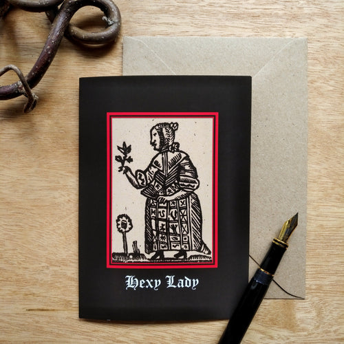Photo of a card featuring a black-and-white mediaeval woodcut artwork on a black background, depicting a woman in a dress and shawl contemplating a plant while holding a folded fan. A flower grows at her feet. Beneath it the caption reads 'Hexy Lady' in a white Gothic typeface. Next to the card is a brown envelope, black fountain pen, and curved piece of wood.