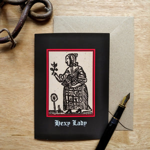 Photo of a card featuring a black-and-white mediaeval woodcut artwork on a black background, depicting a woman in a dress and shawl contemplating a plant while holding a folded fan. A flower grows at her feet. Beneath it the caption reads 'Hexy Lady' in a white Gothic typeface. Next to the card is a brown envelope, black fountain pen, and curved piece of wood.