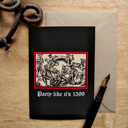 Photo of a card featuring a black-and-white mediaeval woodcut artwork on a black background and framed in red, depicting several demons cavorting around a gigantic ring. One plays some bagpipes, another the lute. Beneath it the caption reads 'Party like it’s 1599' in a white Gothic typeface. Next to the card is a brown envelope, black fountain pen, and curved piece of wood.