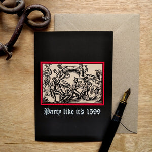 Photo of a card featuring a black-and-white mediaeval woodcut artwork on a black background and framed in red, depicting several demons cavorting around a gigantic ring. One plays some bagpipes, another the lute. Beneath it the caption reads 'Party like it’s 1599' in a white Gothic typeface. Next to the card is a brown envelope, black fountain pen, and curved piece of wood.
