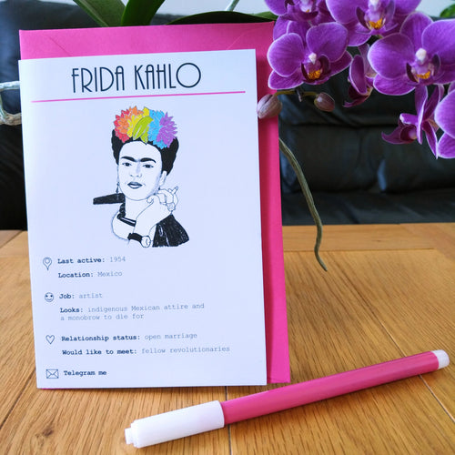 Photo of the card standing in front of a pink envelope. The illustration shows Frida Kahlo on a white background, her hand raised holding a cigarette and her black hair dyed in the colours of the rainbow. The card is titled “Frida Kahlo” and shows numerous details and statuses in line with a modern dating profile. Next to the card is a felt tip pen and some purple flowers.