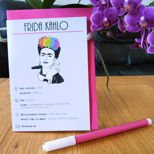 Photo of the card standing in front of a pink envelope. The illustration shows Frida Kahlo on a white background, her hand raised holding a cigarette and her black hair dyed in the colours of the rainbow. The card is titled “Frida Kahlo” and shows numerous details and statuses in line with a modern dating profile. Next to the card is a felt tip pen and some purple flowers.