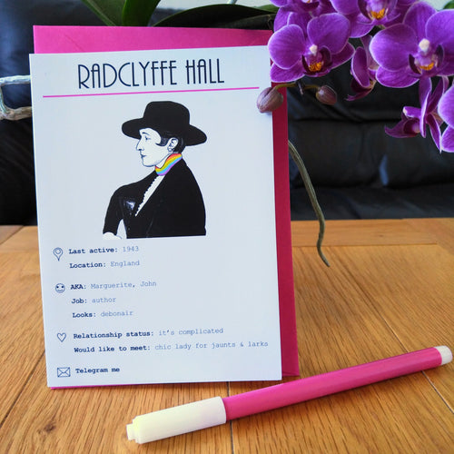 Photo of the card standing in front of a pink envelope. The illustration shows Radyclyffe Hall on a white background, facing to the left and wearing a hat, her scarf in the colours of the rainbow. The card is titled “Radclyffe Hall” and shows numerous details and statuses in line with a modern dating profile. Next to the card is a felt tip pen and some purple flowers.