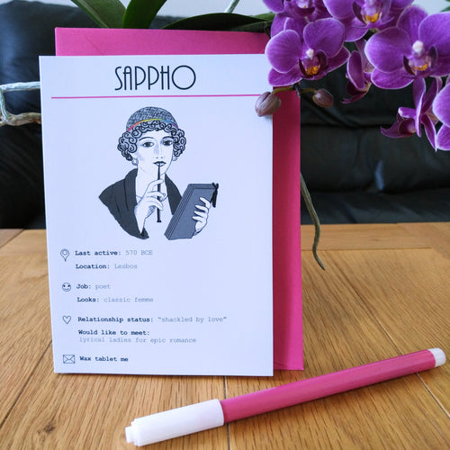 Photo of the card standing in front of a pink envelope. The illustration shows Sappho on a white background. She holds a stylus to her mouth while thinking and holding a wax tablet. A band in her hair is the colours of the rainbow. The card is titled “Sappho” and shows numerous details and statuses in line with a modern dating profile. Next to the card is a felt tip pen and some purple flowers.