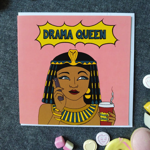 Photo of a large square pink greetings card featuring a comic book style illustration of Cleopatra on her mobile phone and holding a take-away coffee, with 'Drama Queen' written in a flash above her. Dotted around the card & white envelope are sweets in pinks, yellows and whites to match the card colours.
