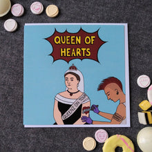Load image into Gallery viewer, Photo of a light blue card featuring Queen Victoria, seated, wearing a black dress, white headdress, and coronet, along with a sash that reads “Queenz On Tour”. She is receiving a tattoo on her left arm of a red heart with “Albert” across it in a banner. The tattooist has a red Mohican, earrings, and a “YAASSS QUEEN” tattoo. The main caption reads “Queen Of Hearts” in yellow text in a dark red exploding bubble. Next to the card can be found some sweets and biscuits.