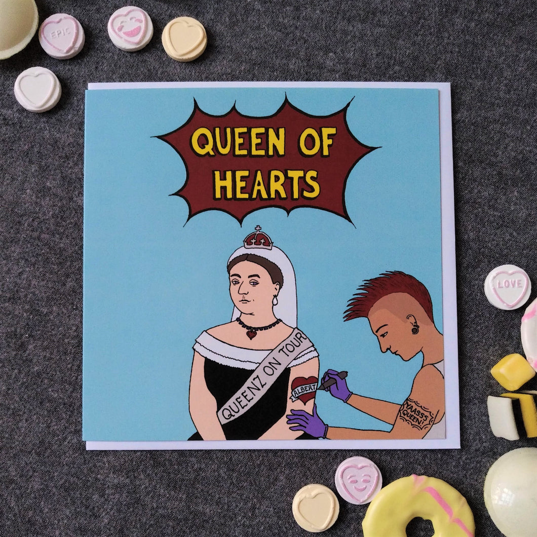 Photo of a light blue card featuring Queen Victoria, seated, wearing a black dress, white headdress, and coronet, along with a sash that reads “Queenz On Tour”. She is receiving a tattoo on her left arm of a red heart with “Albert” across it in a banner. The tattooist has a red Mohican, earrings, and a “YAASSS QUEEN” tattoo. The main caption reads “Queen Of Hearts” in yellow text in a dark red exploding bubble. Next to the card can be found some sweets and biscuits.