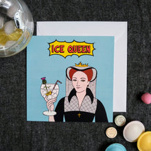 Load image into Gallery viewer, Photo of a light blue card featuring Mary Queen of Scots smirking and toasting with a large cocktail in a glass, with ice, lemon, a straw, and a flower. The caption reads “Ice Queen” in red text inside a yellow exploding bubble. Next to the card are some sweets and a drink with ice.