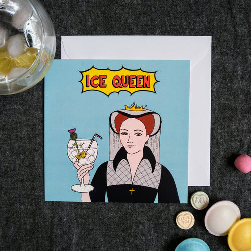 Photo of a light blue card featuring Mary Queen of Scots smirking and toasting with a large cocktail in a glass, with ice, lemon, a straw, and a flower. The caption reads “Ice Queen” in red text inside a yellow exploding bubble. Next to the card are some sweets and a drink with ice.