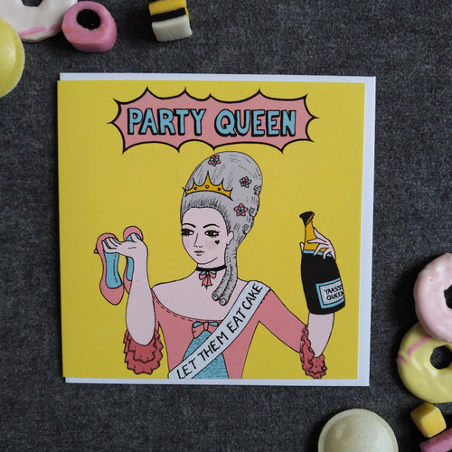 Photo of a light yellow card featuring Marie Antoinette with large silver hair and a crown, wearing a blue and pink dress and a sash that reads “Let Them Eat Cake”. She holds a pair of pink high hells over her shoulder and holds an opened champagne bottle in the other hand. The caption reads “Party Queen” in blue text inside a pink exploding bubble. Next to the card are some sweets and biscuits.