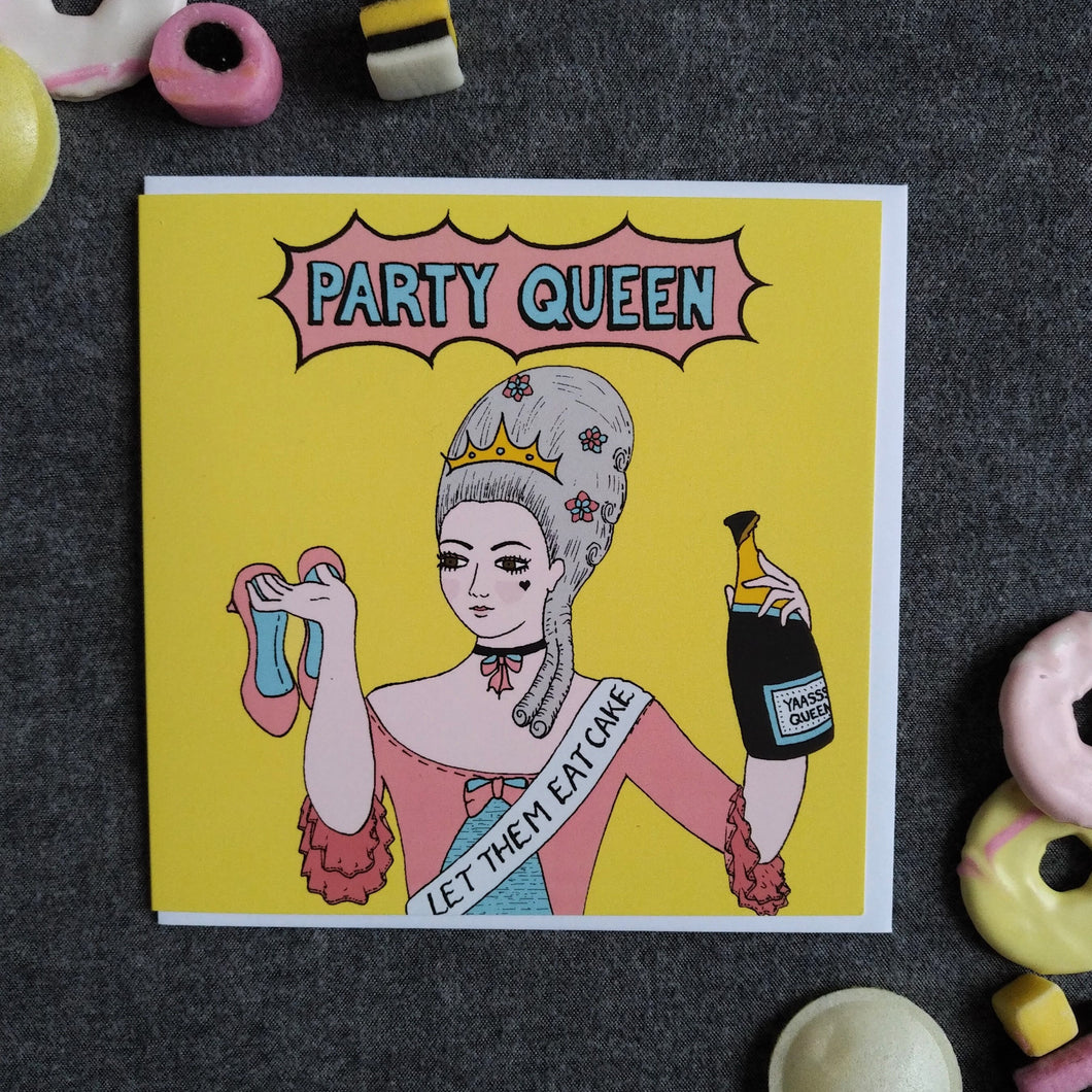 Photo of a light yellow card featuring Marie Antoinette with large silver hair and a crown, wearing a blue and pink dress and a sash that reads “Let Them Eat Cake”. She holds a pair of pink high hells over her shoulder and holds an opened champagne bottle in the other hand. The caption reads “Party Queen” in blue text inside a pink exploding bubble. Next to the card are some sweets and biscuits.