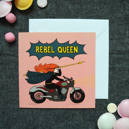 Photo of a pink card featuring Boudicca, with her ginger hair in a whirl around her as she raises a yellow spear and rides a motorcycle. Above her, a dark blue exploding bubble with “Rebel Queen” in yellow text. Next to the card are various sweets.