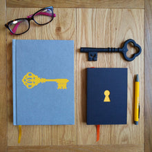 Load image into Gallery viewer, Photograph of the Escape Journal, a black paperback with a yellow keyhole cut-out and orange placeholder ribbon, alongside the Liberation Journal, a yellow pen, a black iron key, and a folded pair of glasses.