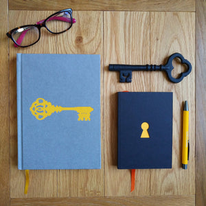 Photograph of the Escape Journal, a black paperback with a yellow keyhole cut-out and orange placeholder ribbon, alongside the Liberation Journal, a yellow pen, a black iron key, and a folded pair of glasses.