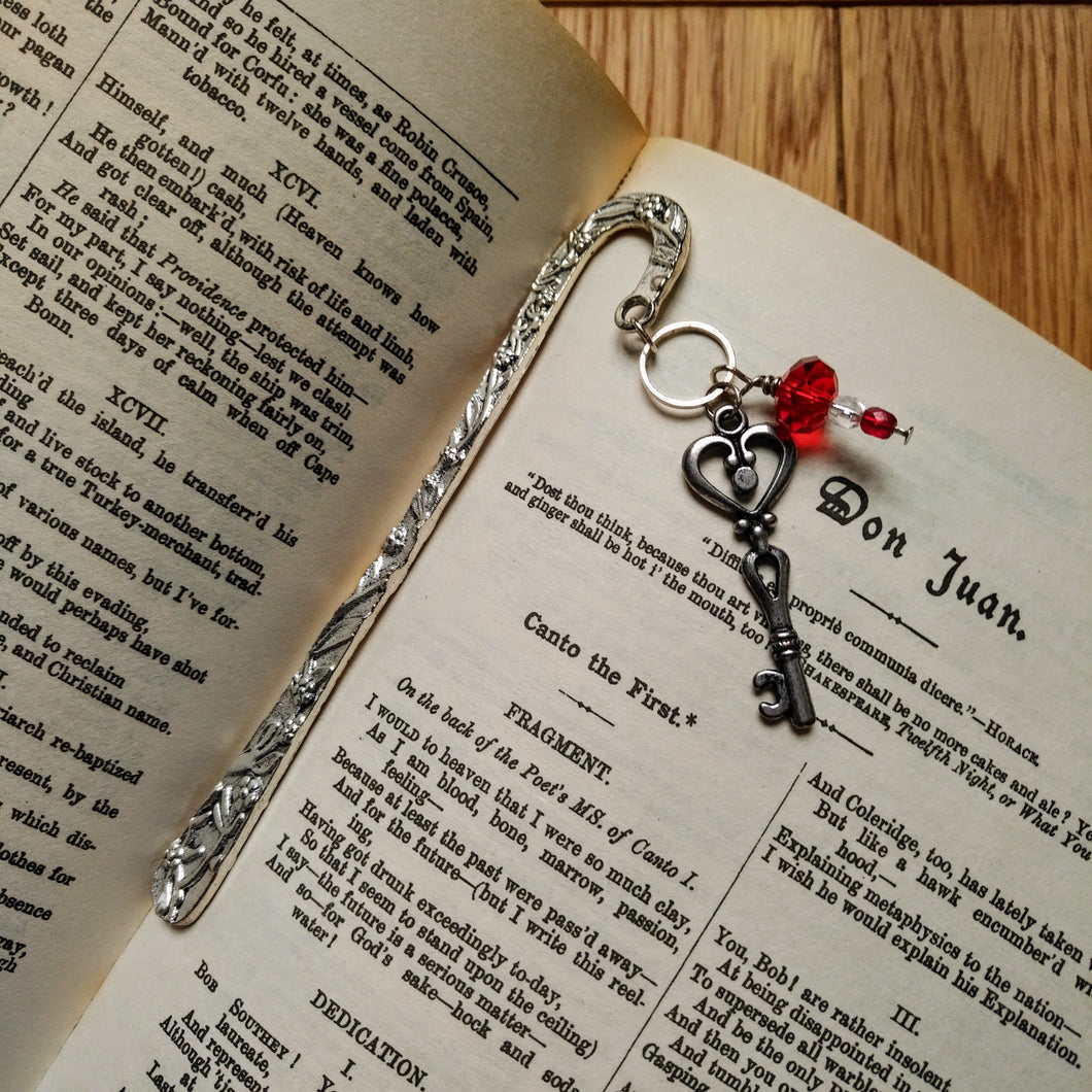 Photo of a silver crook-shaped bookmark that is joined at the tip by a silver key charm with a heart shape in the handle. Crimson beads are also attached. The page saver lies on the page of an open book.