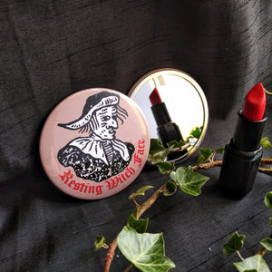 Photo of a two pocket mirrors. One shows the back, with a brown background and black-and-white mediaeval woodcut artwork of stereotypical crone with warts and a large nose. The caption reads 'Resting Witch Face' in a red Gothic typeface. The other mirror reflects a red lipstick and branch with leaves.
