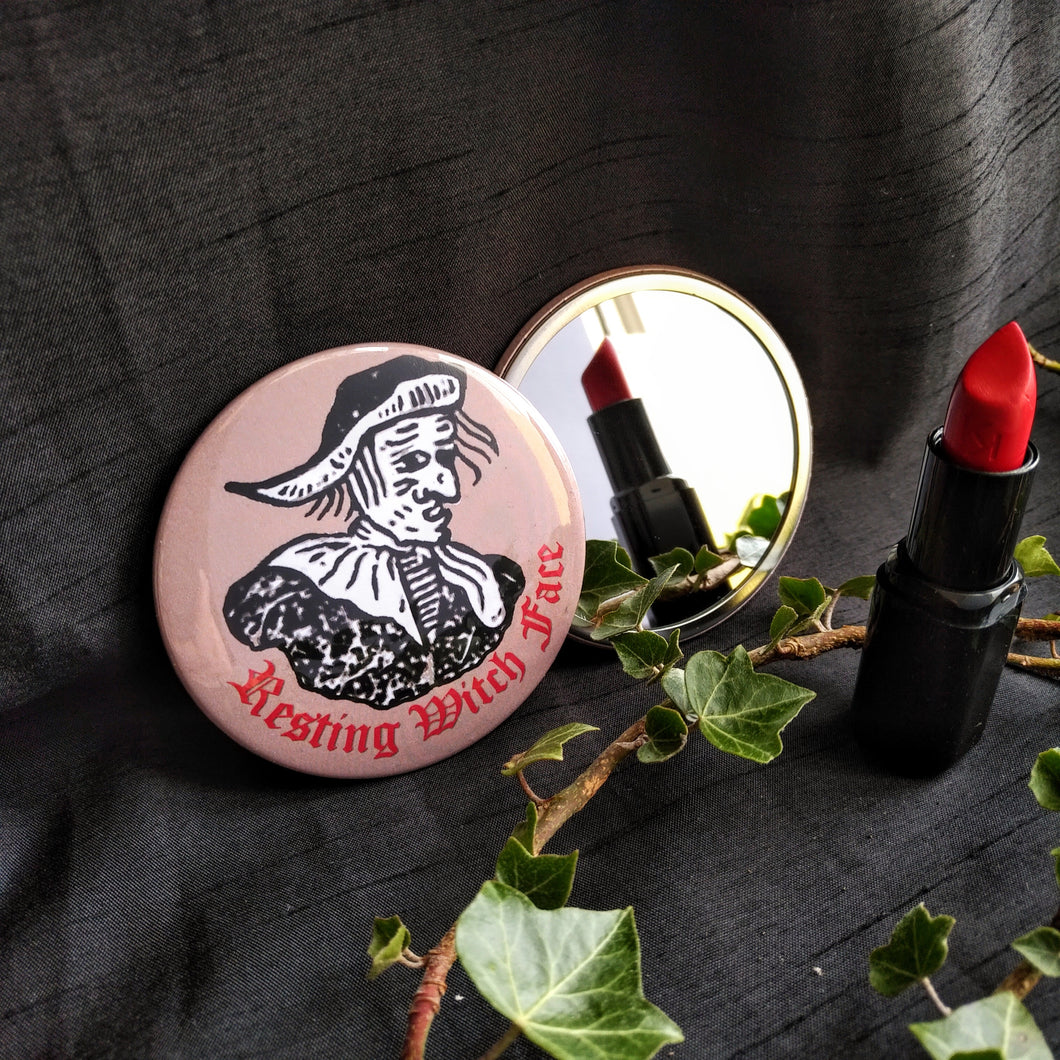 Photo of a two pocket mirrors. One shows the back, with a brown background and black-and-white mediaeval woodcut artwork of stereotypical crone with warts and a large nose. The caption reads 'Resting Witch Face' in a red Gothic typeface. The other mirror reflects a red lipstick and branch with leaves.