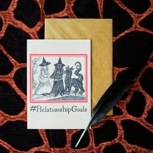 A buff coloured card featuring a mediaeval woodcut artwork depicting two witches, one of which is holding a pet demon. Beneath it the caption reads #RelationshipGoals. Pictured with a brown envelope, black feather and black pattered background.